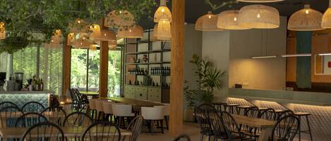 Un restaurant doté de nombreuses tables et chaises, de luminaires suspendus en fibres tressées et offrant une vue sur la verdure environnante.