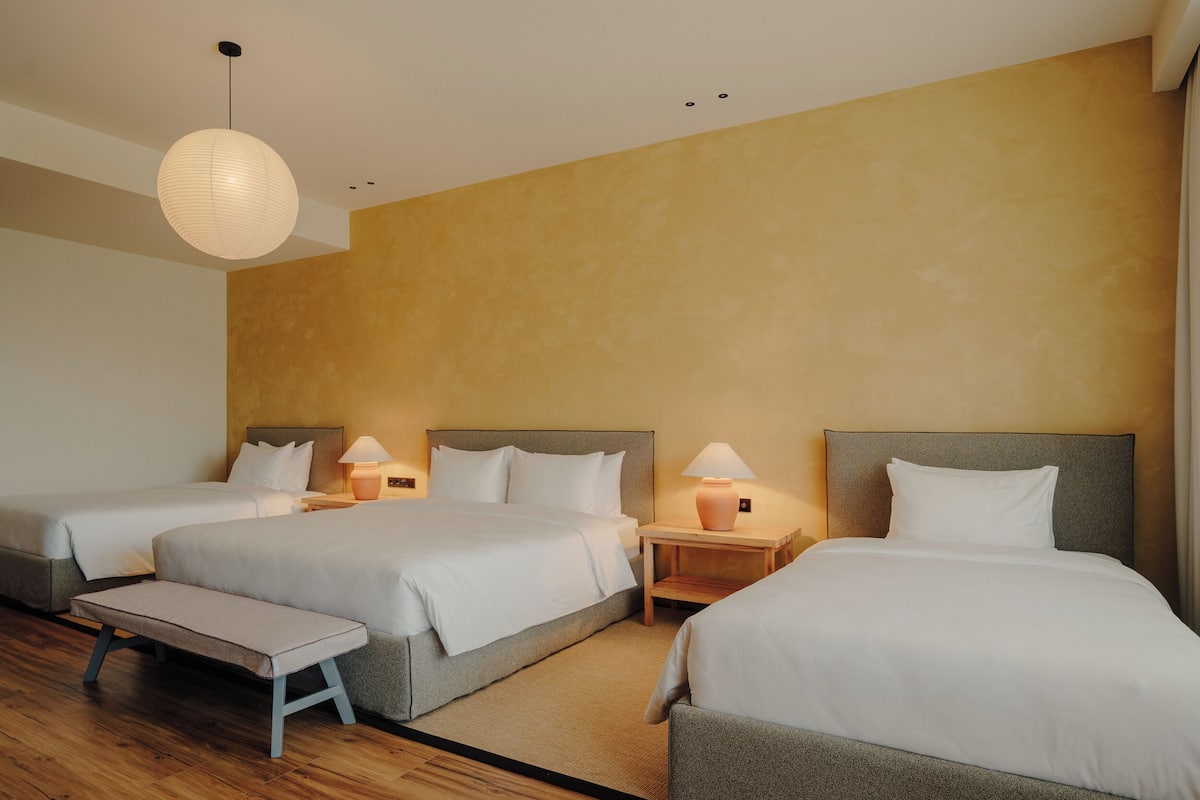 A hotel room with three beds, a bench, and a large wall-mounted light fixture.
