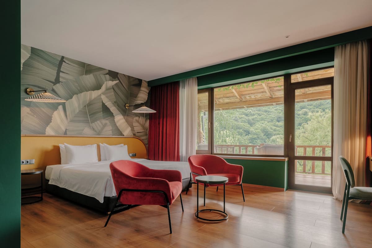 A hotel room with a bed, two red chairs, a small table, a ceiling fan, and a large window with a view of greenery.