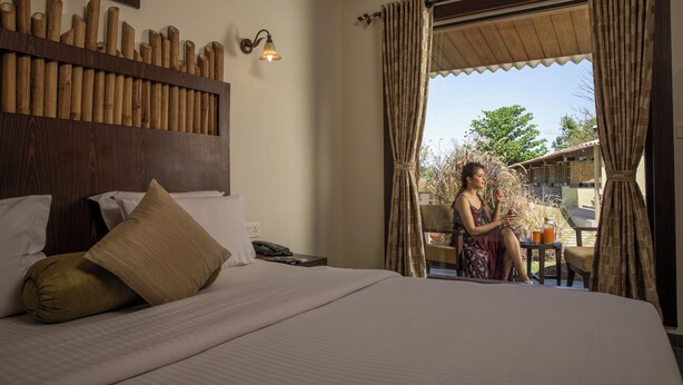 A hotel room with a large bed, a wooden headboard, a wall-mounted light fixture, and a view of the outdoors through a window with curtains. - Asiatic Lion Lodge (Mendarda)