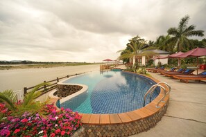 A swimming pool with a wooden deck and lounge chairs, surrounded by tropical plants and palm trees. - Myitkyina Plam Spring Resort Hotel (Myitkyina)