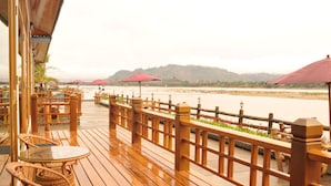 A wooden deck with tables and chairs, overlooking a river with a wooden railing and red umbrellas. - Myitkyina Plam Spring Resort Hotel (Myitkyina)