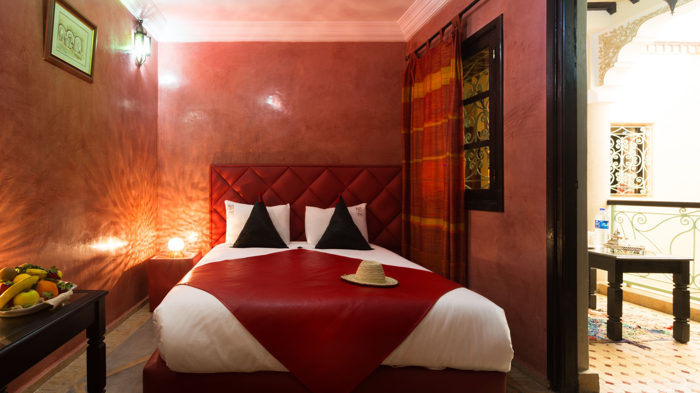A hotel room with a red and white bed, a nightstand with fruit, and a view of the outside balcony.