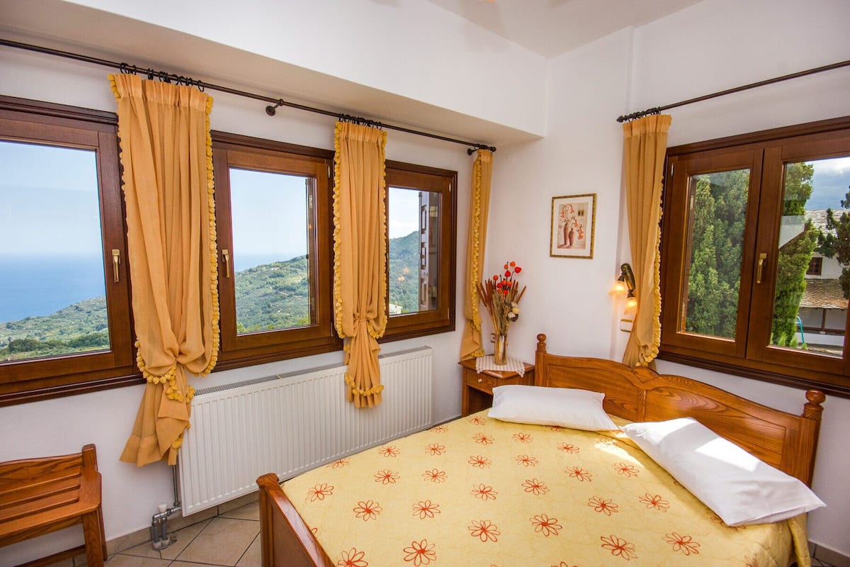 A bedroom with a bed, a window with curtains, a radiator, a bedside table with a lamp, and a view of the sea.