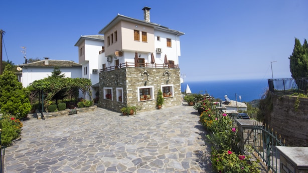 A stone-paved courtyard with a two-story building, a stone wall, and a view of the sea. - Guesthouse Papagiannopoulou (Zagora-Mouresi)