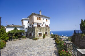 A stone-paved courtyard with a two-story building, a stone wall, and a view of the sea. - Guesthouse Papagiannopoulou (Zagora-Mouresi)