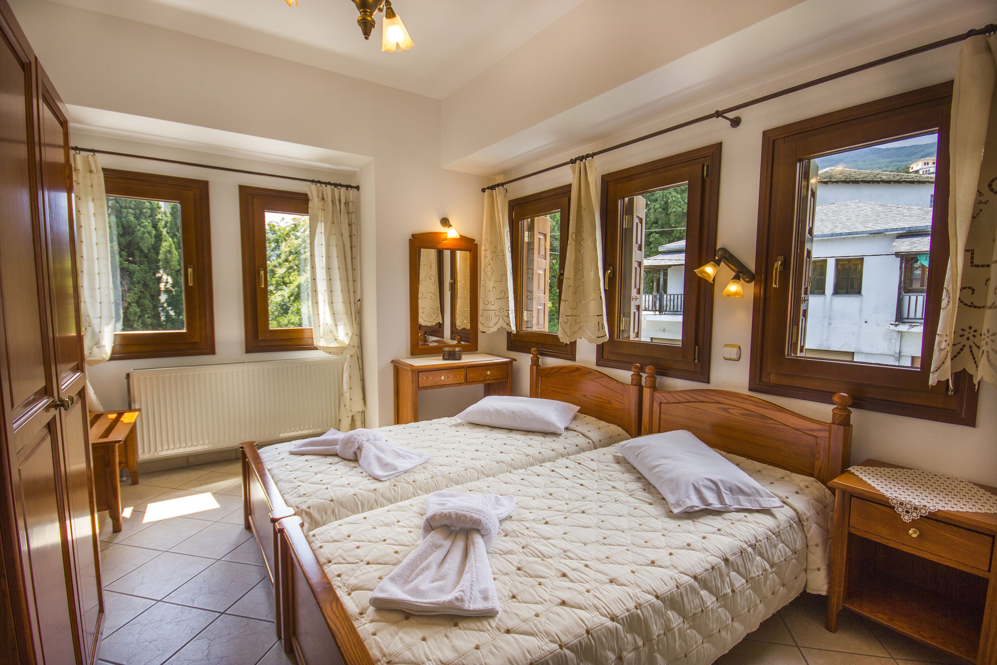 A hotel room with two beds, a wooden headboard, a bedside table with a lamp, and a view of the outside through large windows.