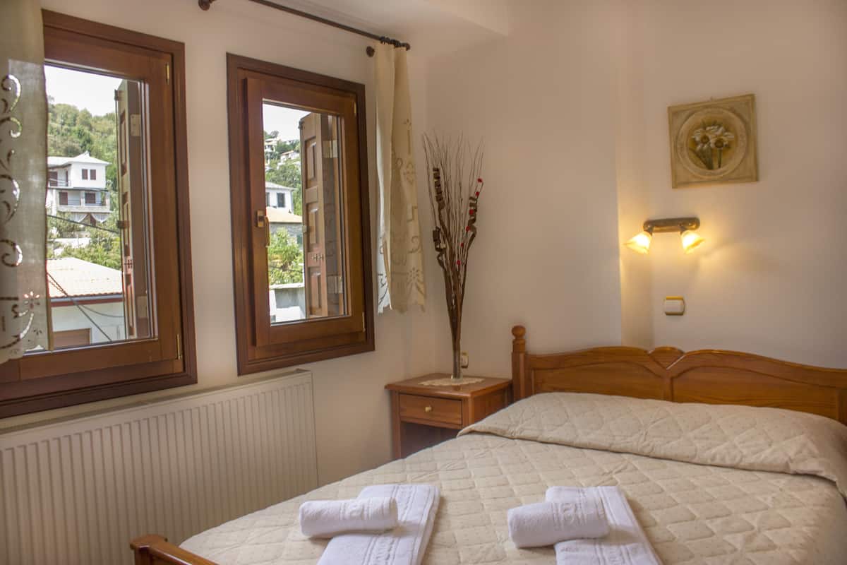 A bedroom with two beds, a window with a view of buildings, a nightstand with a vase, and a wall-mounted light fixture.