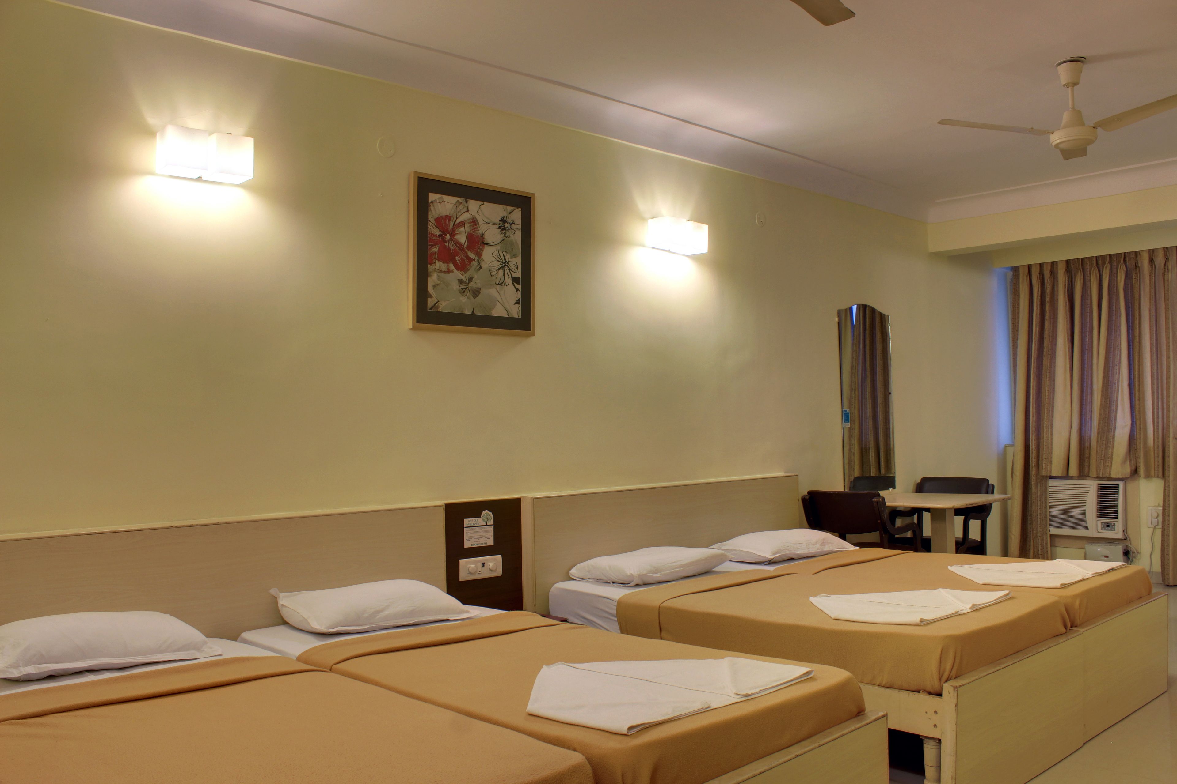 Habitació d'hotel amb tres llits individuals, un escriptori, una cadira, un ventilador de sostre i un aire condicionat fixat a la paret.