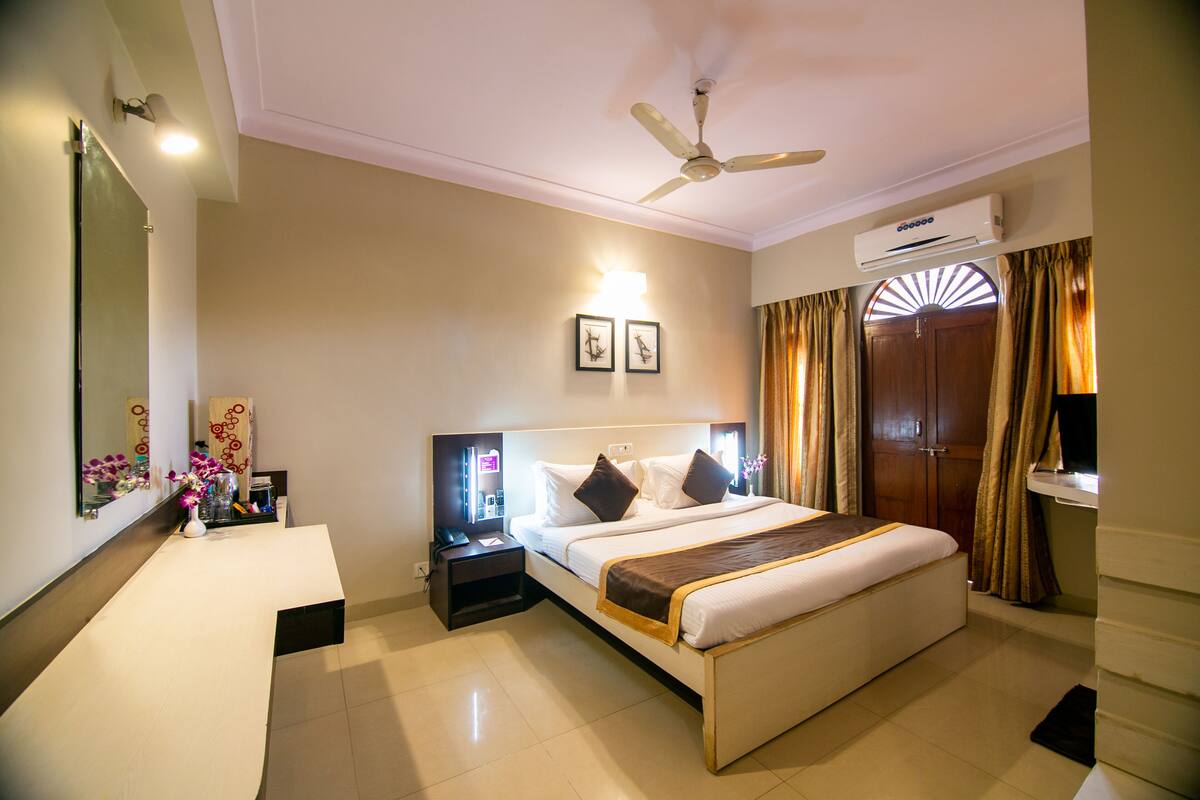 A hotel room with a large bed, a bedside table, a nightstand, a mirror, a ceiling fan, and an air conditioning unit.