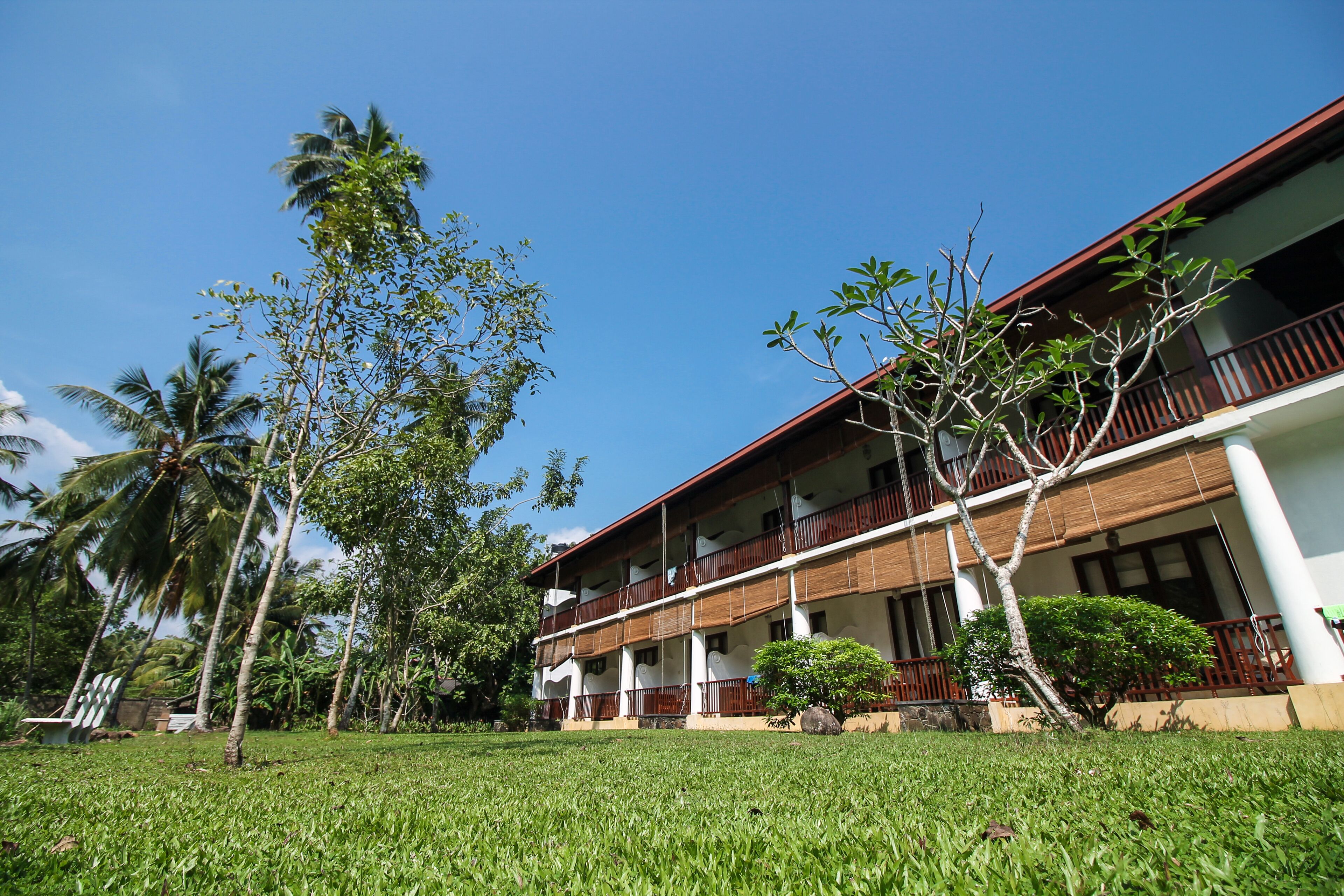 A two-story building with a balcony on each floor, surrounded by greenery and palm trees.