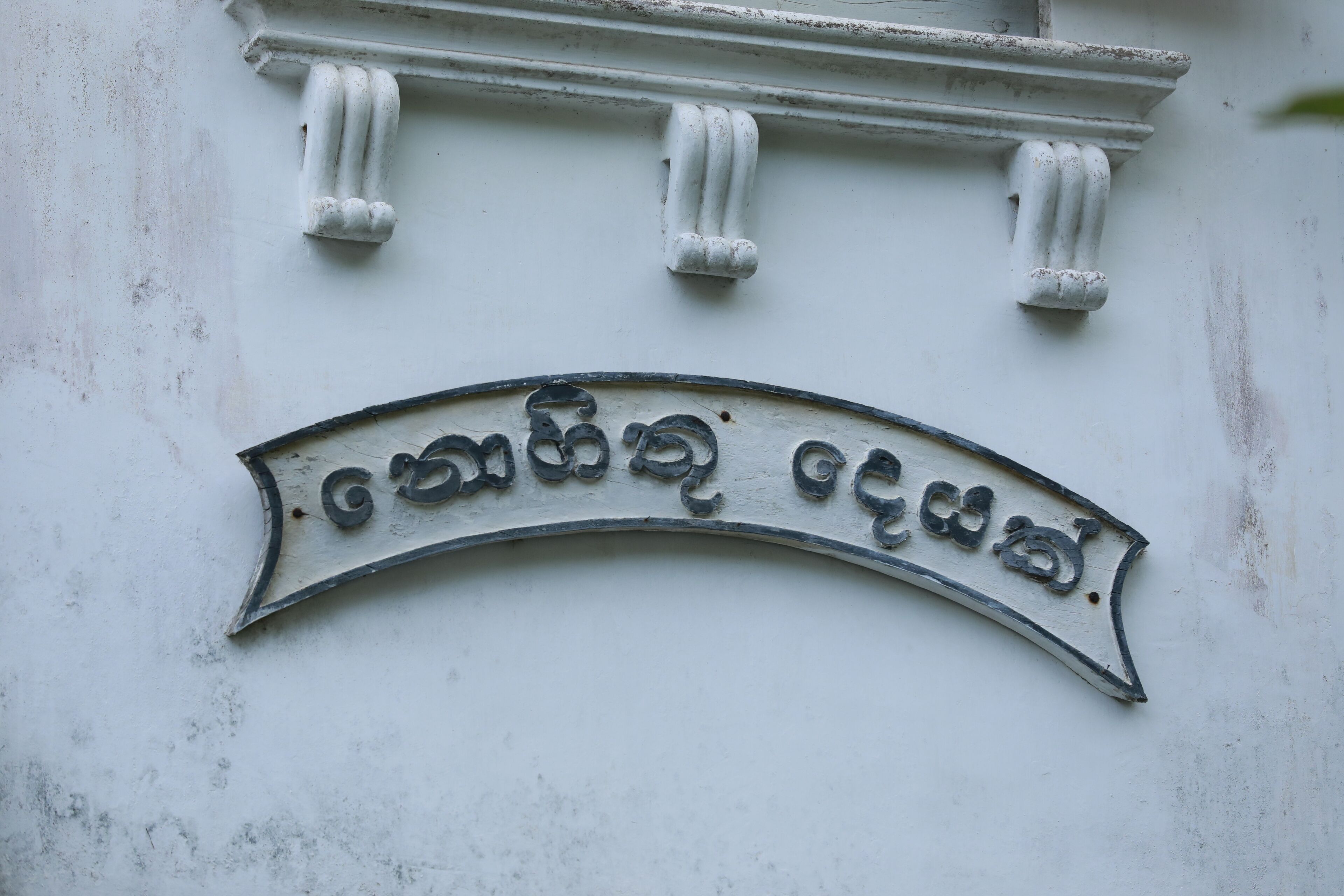 A white wall with a curved metal sign featuring text in a non-Latin script, likely indicating a location or name.
