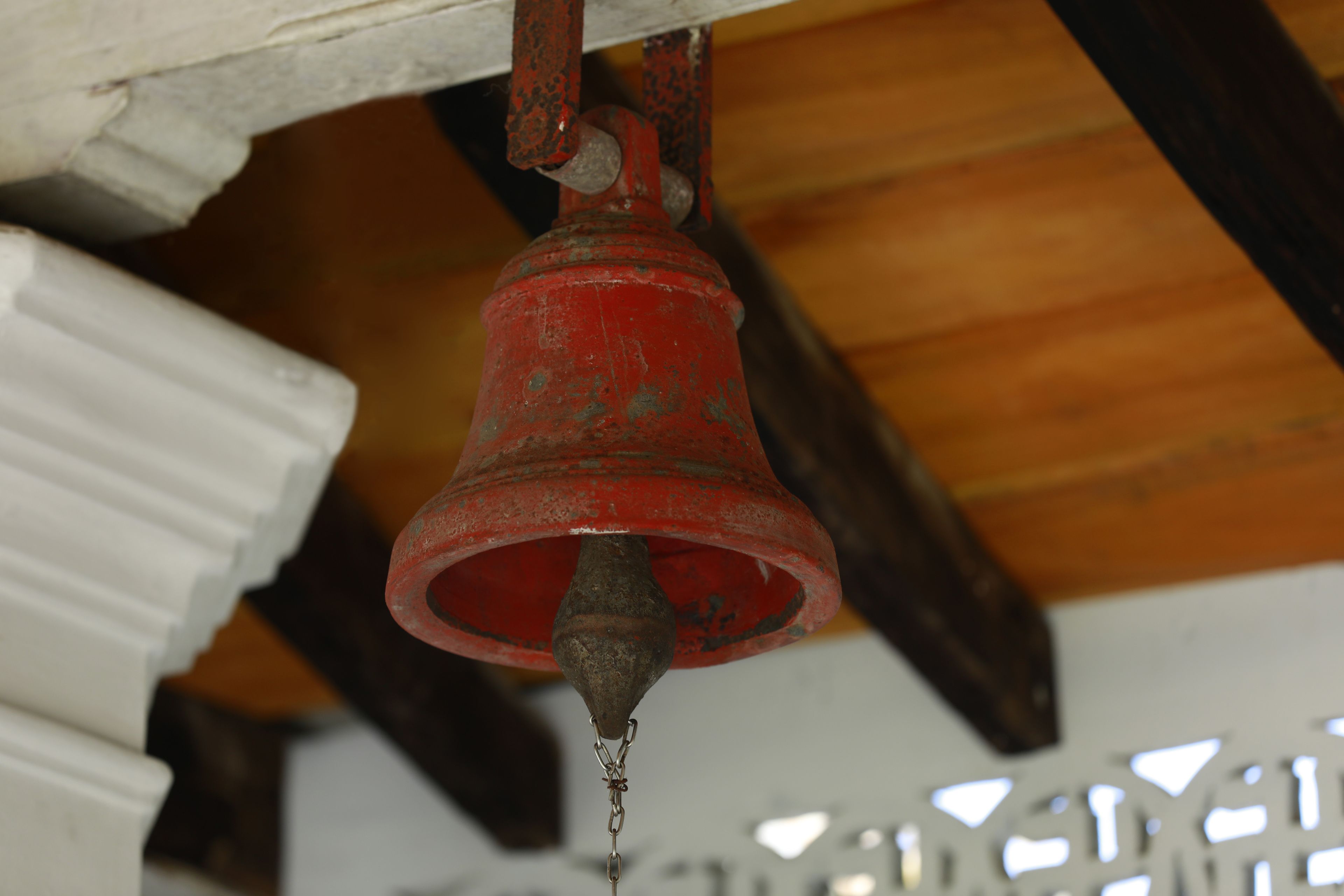 A red bell hanging from a wooden ceiling, attached to a white column.