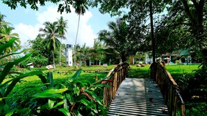 A bamboo bridge in a lush garden with palm trees and a white building in the background. - Nooit Gedacht Heritage Hotel-Original Dutch Governor's House (Unawatuna)