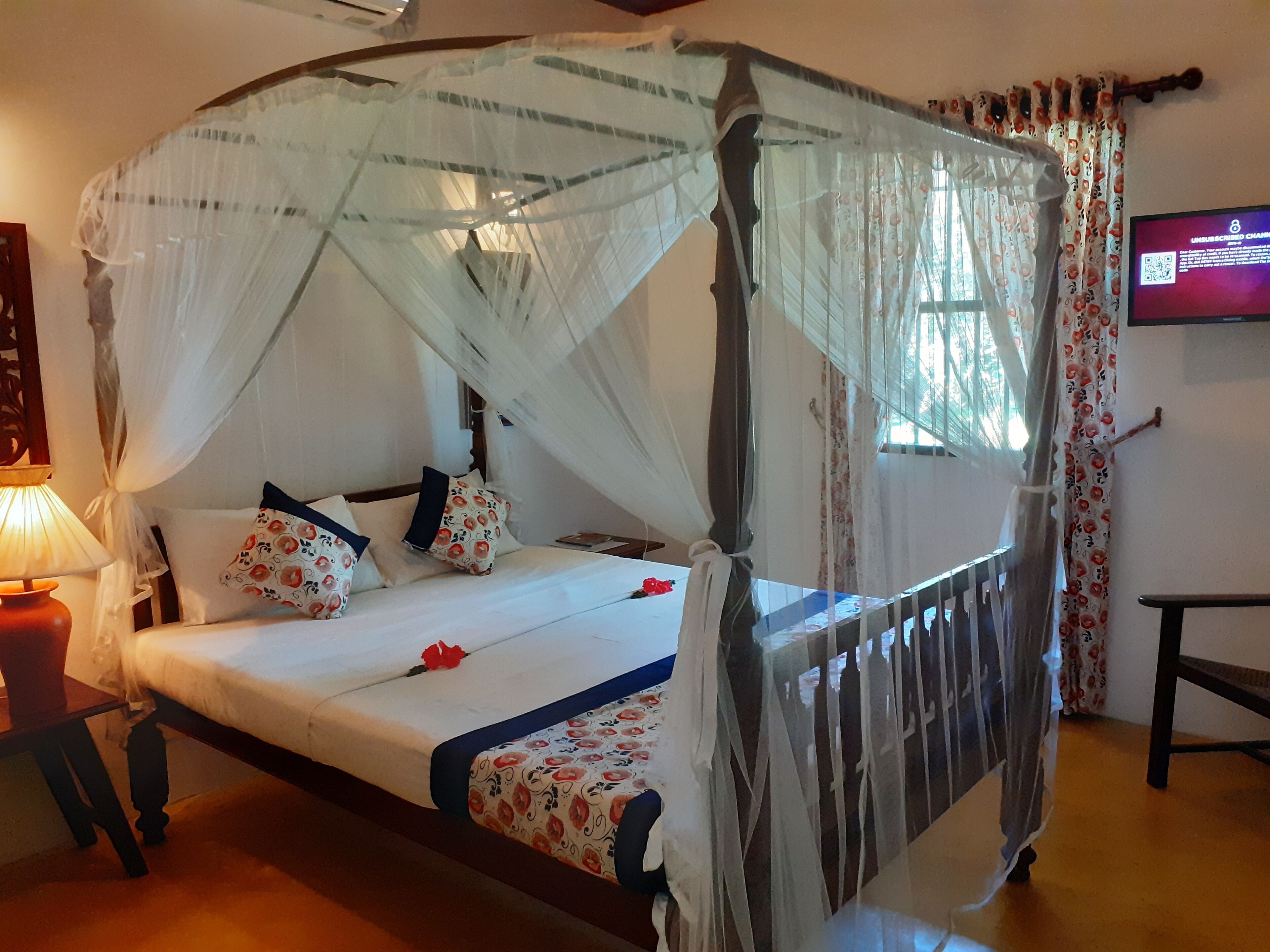 A four-poster bed with a canopy, a bedside table with a lamp, and a television on the wall.