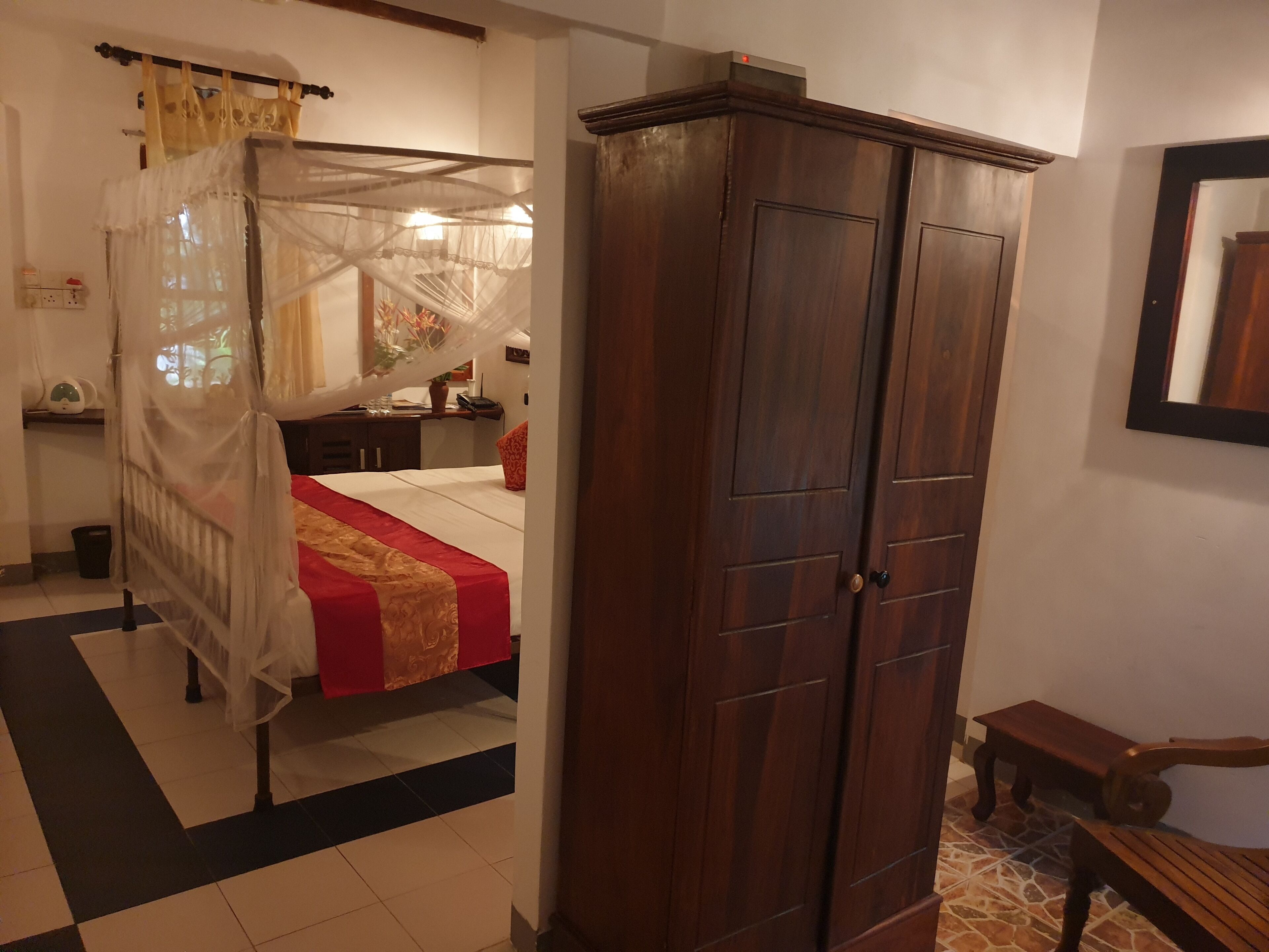 A bedroom with a four-poster bed, a wooden wardrobe, a bench, and a tiled floor.