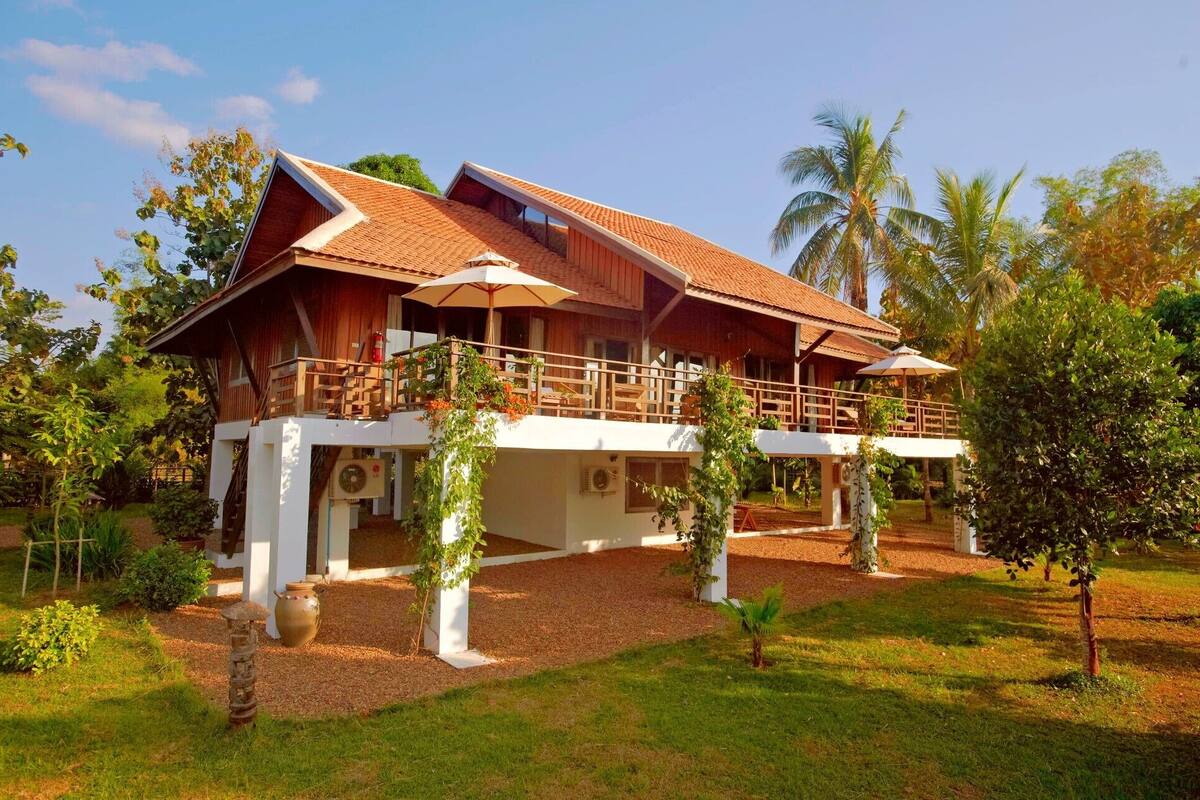 A two-story wooden house with a balcony, surrounded by lush greenery and palm trees.