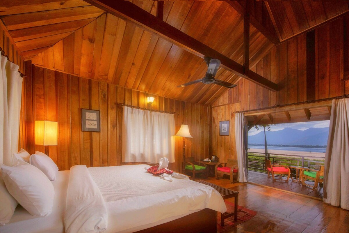 A wooden cabin bedroom with a bed, two bedside lamps, a ceiling fan, and a balcony with chairs and a table.