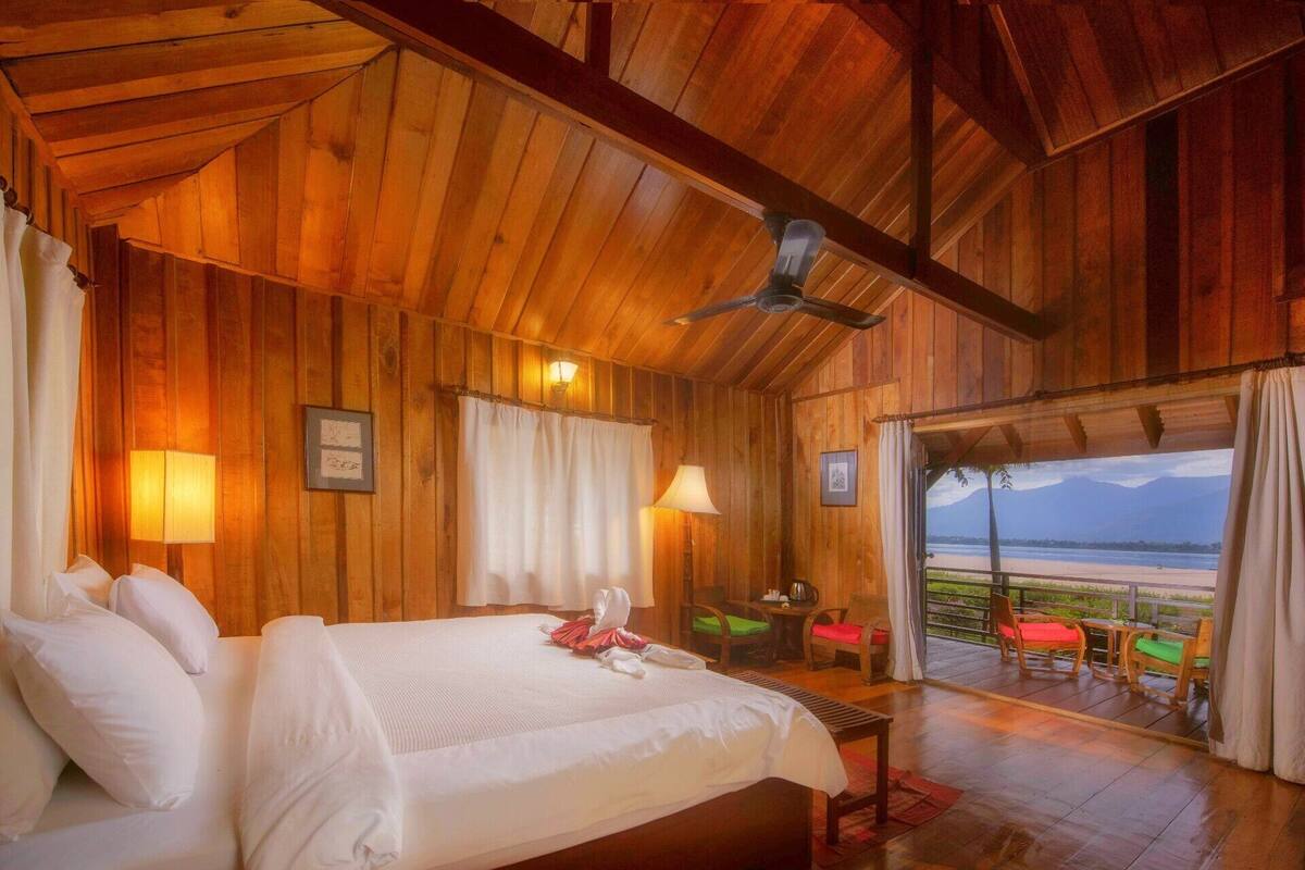 A wooden cabin bedroom with a bed, two bedside lamps, a ceiling fan, and a balcony with chairs and a table.