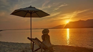Escena de playa con una persona sentada bajo una sombrilla, una silla de playa y una puesta de sol sobre el agua.