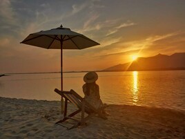 Escena de playa con una persona sentada bajo una sombrilla, una silla de playa y una puesta de sol sobre el agua.