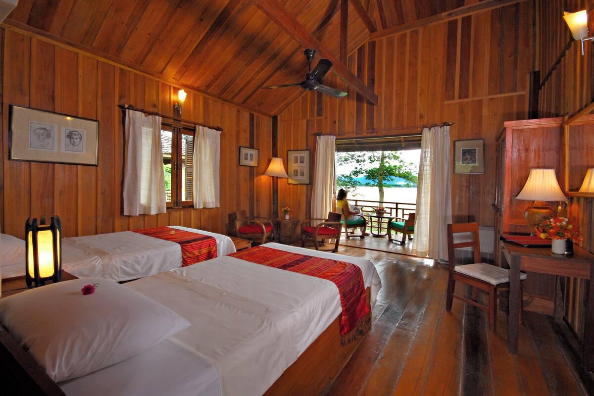 A room with wooden walls, two beds with white linens, a ceiling fan, a table with chairs, and a view through a window.