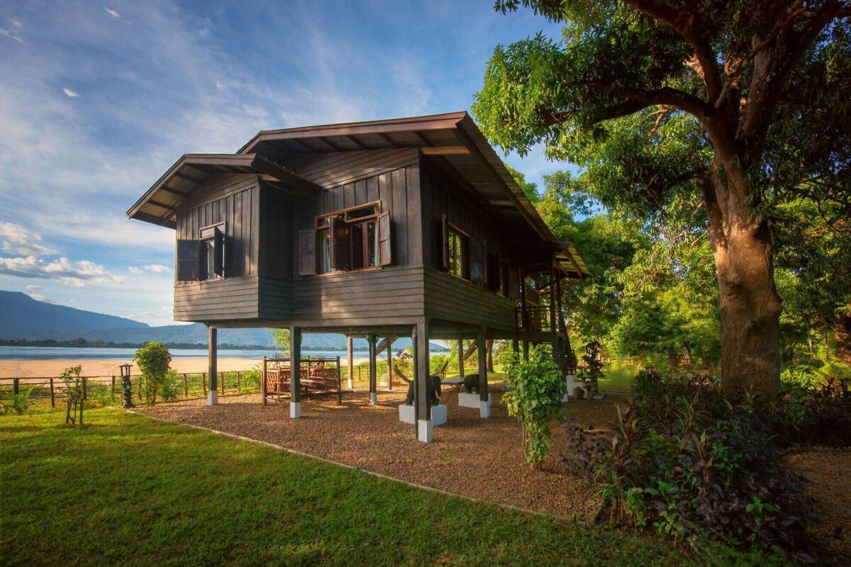 A wooden house with a balcony, situated near a body of water and surrounded by greenery.