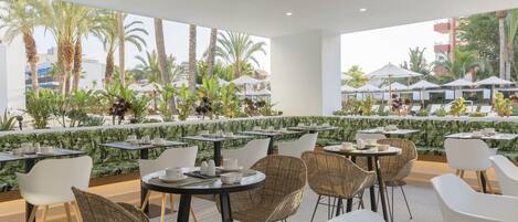 Un área de comedor al aire libre con mesas y sillas, rodeada de follaje y palmeras.