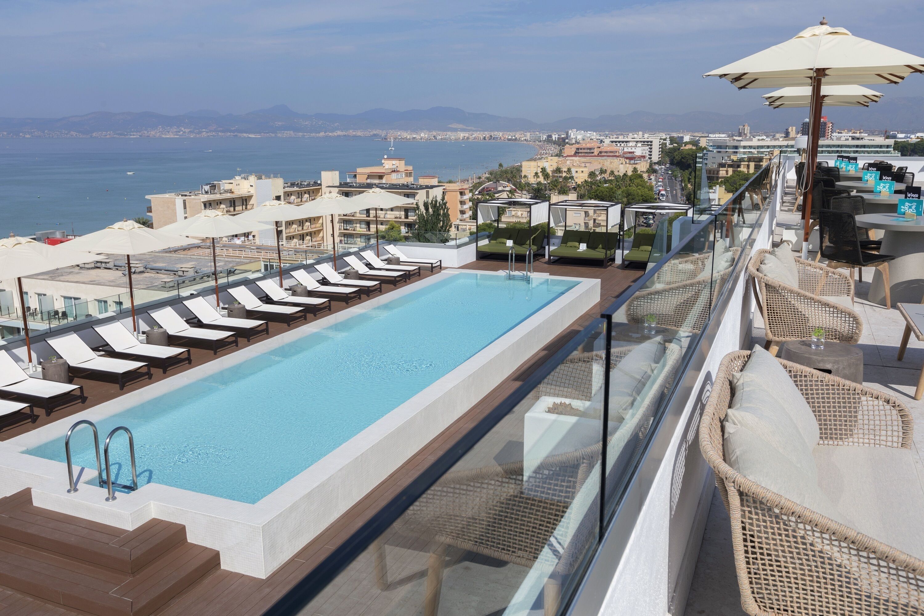 A rooftop pool with lounge chairs and umbrellas, overlooking a cityscape and the sea.