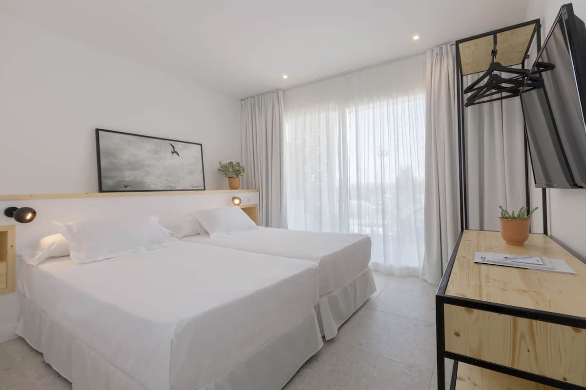 A hotel room with a bed, bedside tables, a desk with a chair, a potted plant, a wall-mounted TV, and a window with curtains.