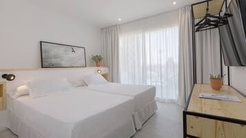 Una habitación de hotel con una cama, mesitas de noche, un escritorio con silla, una planta en maceta, un televisor montado en la pared y una ventana con cortinas.