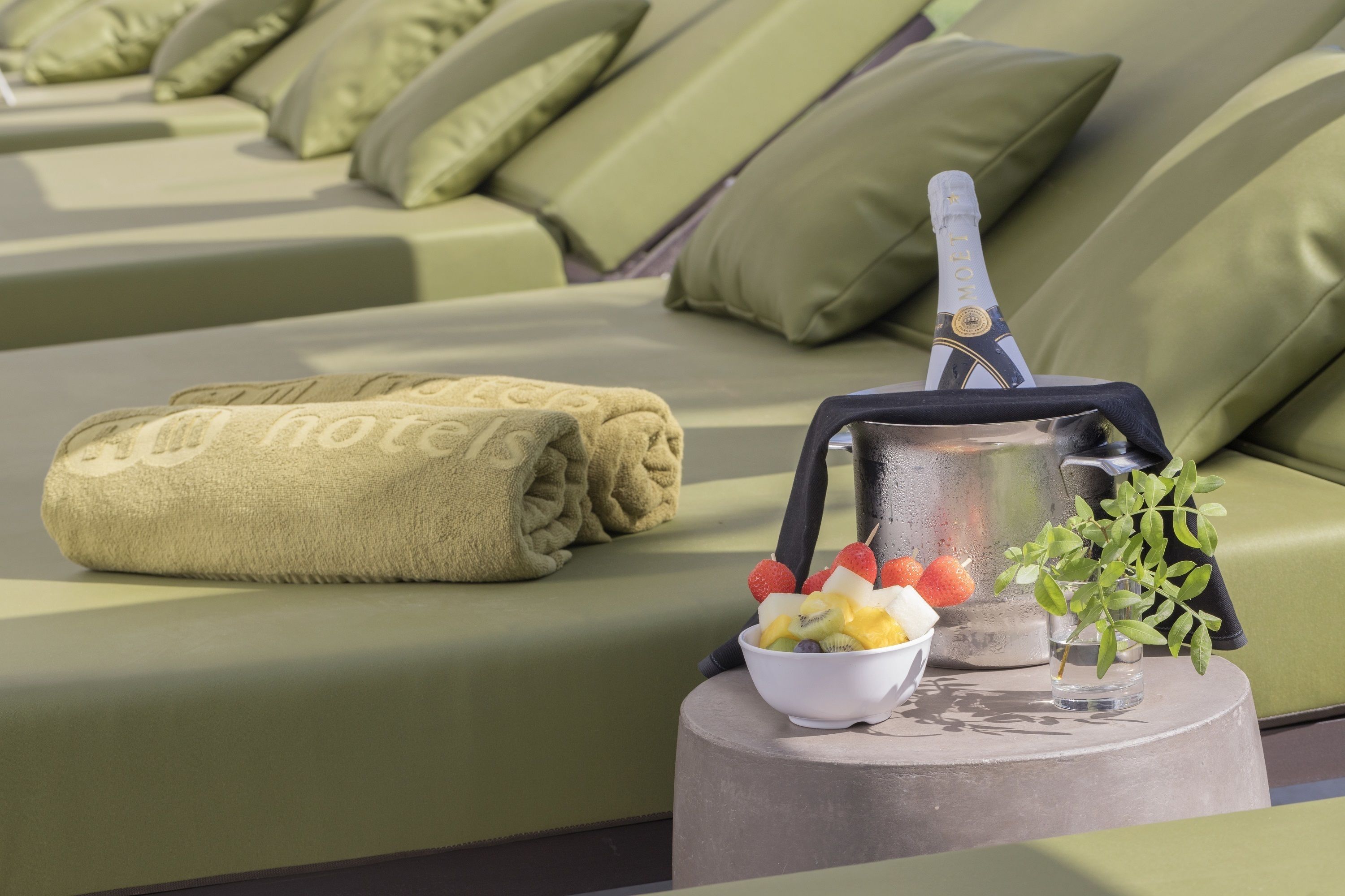 A sun lounger with a towel, a bowl of fruit, a glass of water, and a bottle of champagne.