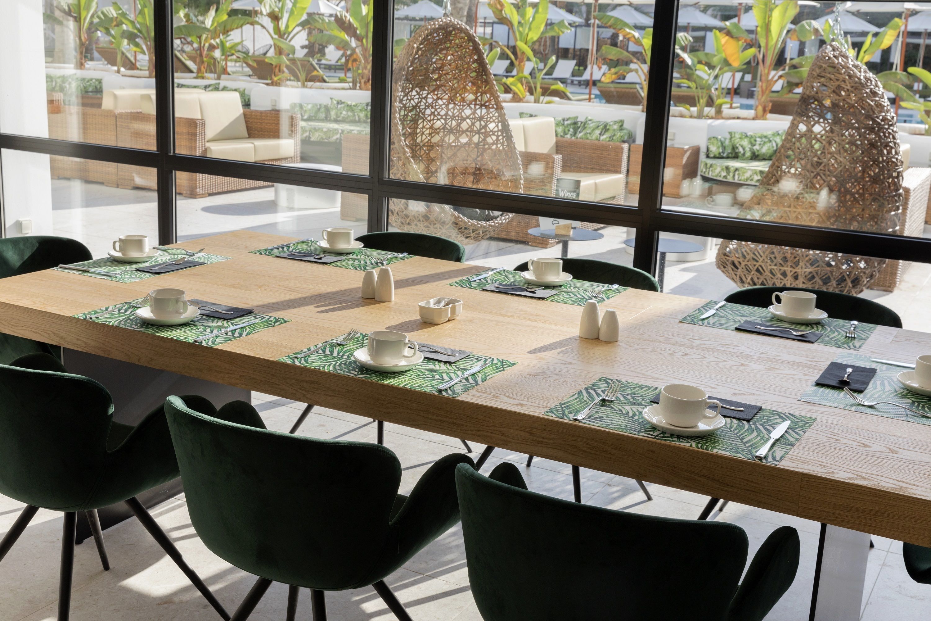 Comedor con una larga mesa dispuesta para seis personas, rodeada de sillas verdes, con vista a un área de estar exterior y plantas a través de amplios ventanales.