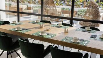 Comedor con una larga mesa dispuesta para seis personas, rodeada de sillas verdes, con vista a un área de estar exterior y plantas a través de amplios ventanales.