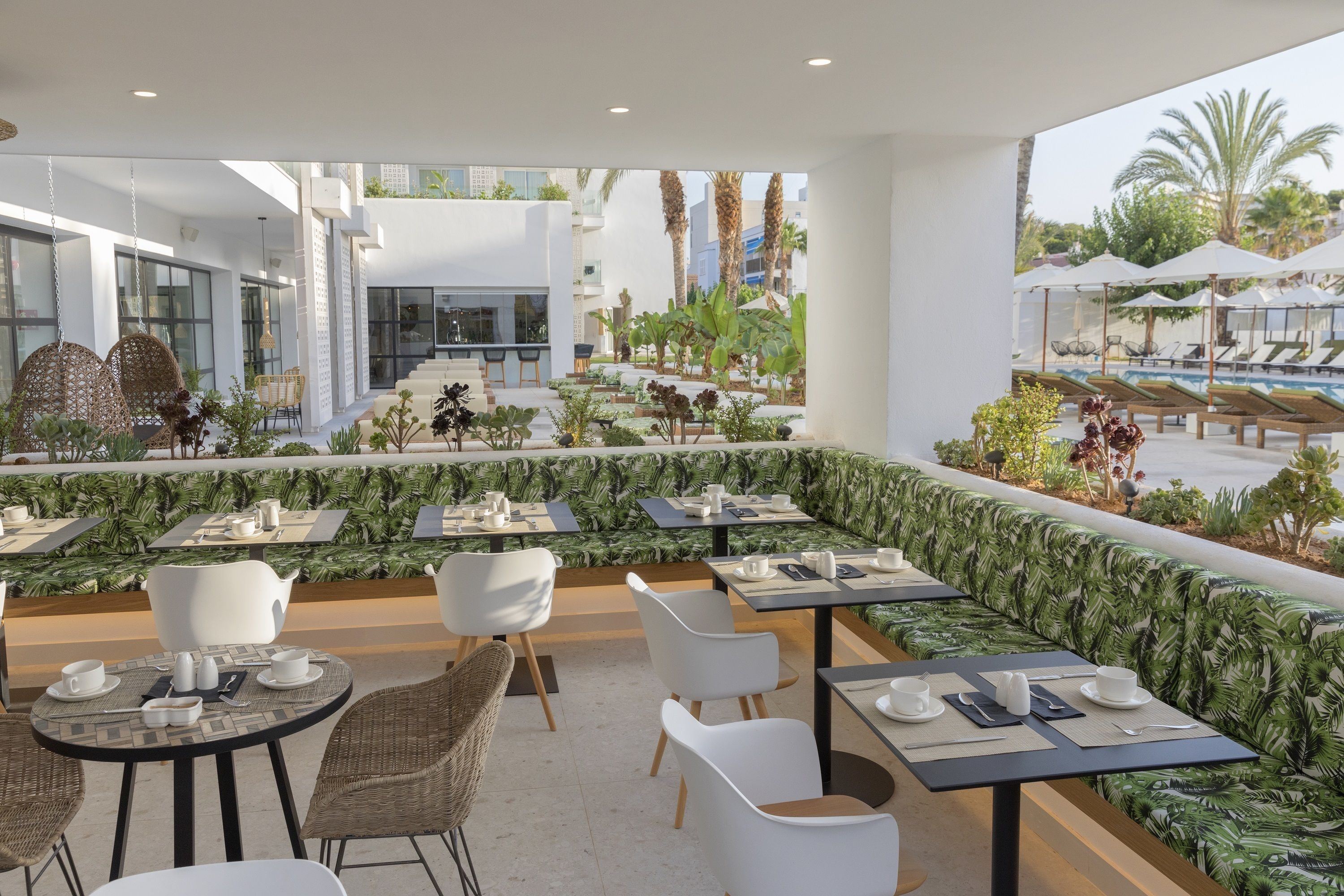 Un área de comedor en la azotea con mesas y sillas, una piscina al fondo y vistas a palmeras y edificios.