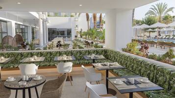 Un área de comedor en la azotea con mesas y sillas, una piscina al fondo y vistas a palmeras y edificios.