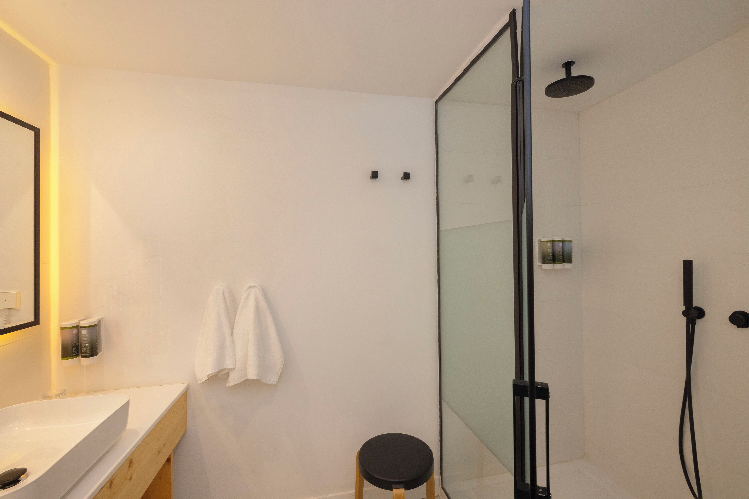 Un baño moderno con cabina de ducha de vidrio, un lavabo blanco y un taburete de madera.