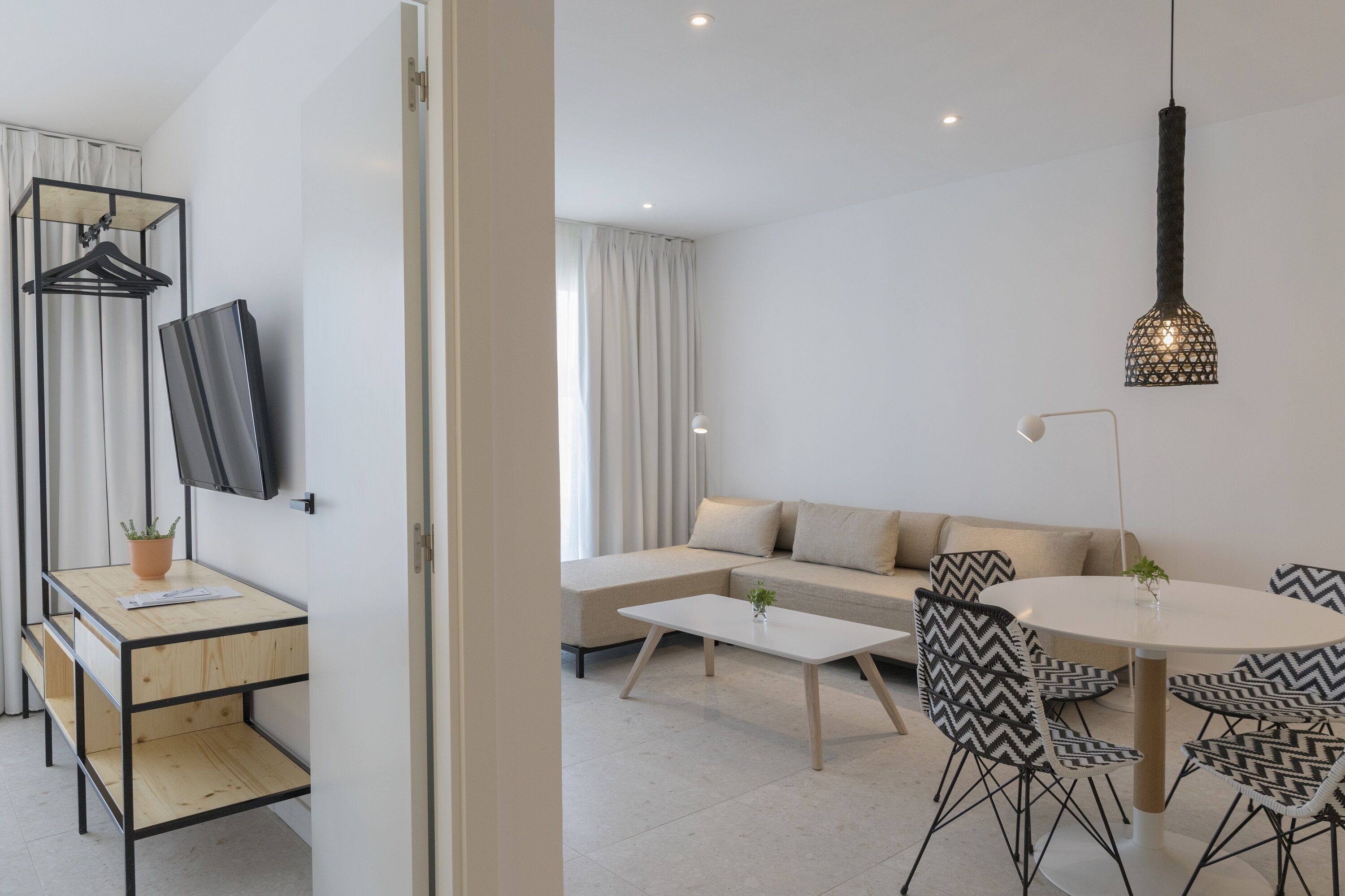 Een moderne woonkamer met een bank, een salontafel en een eettafel met stoelen. Er is een televisie aan de muur bevestigd en een hanglamp.
