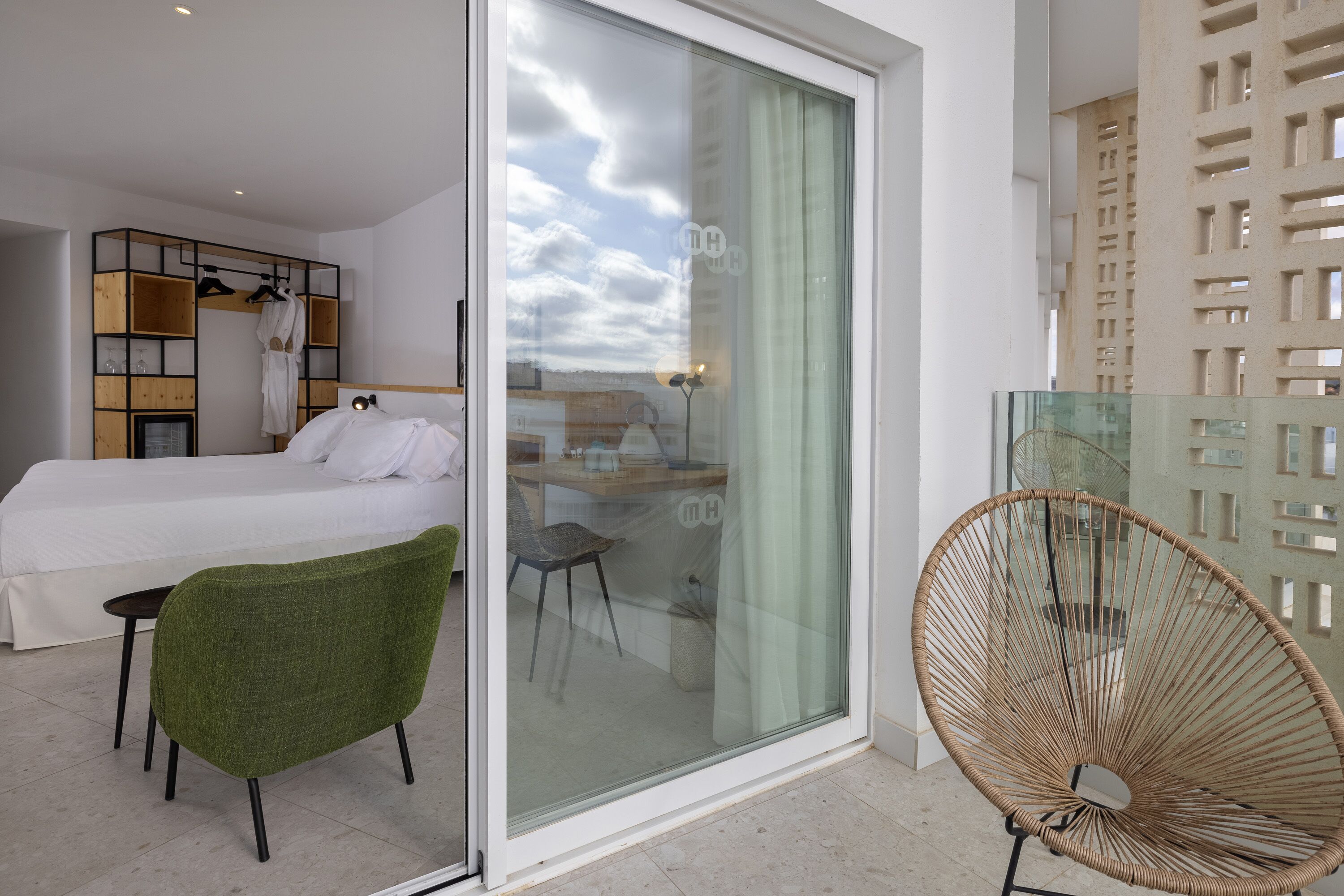 A modern hotel room with a bed, a green chair, a glass table, and a glass door leading to a balcony.