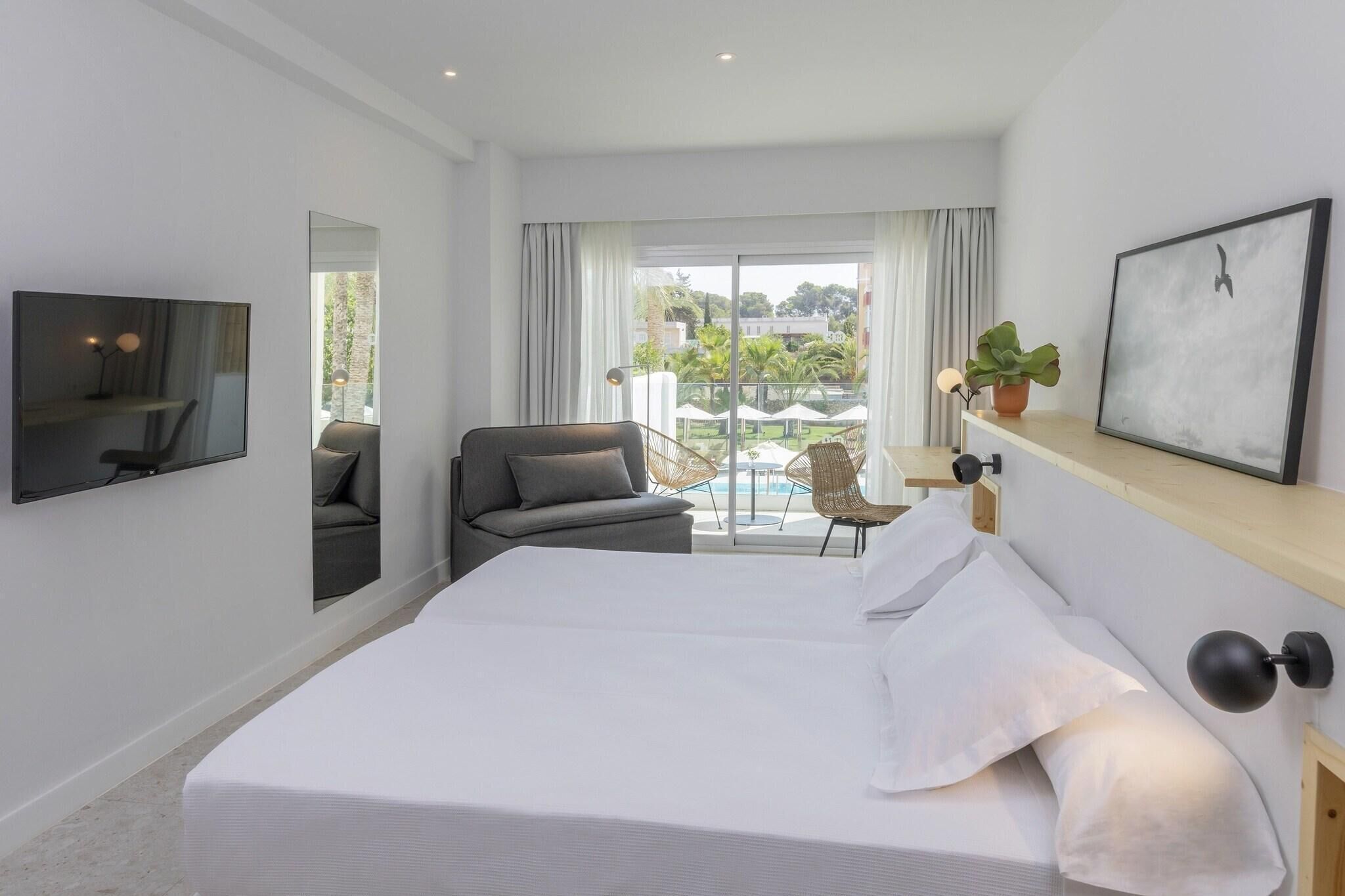 A hotel room with a bed, a TV, a sofa, a chair, a plant, and a balcony with a view.