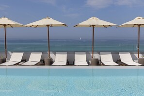 Zona junto a la piscina con sillones de descanso y sombrillas, con vista al océano.