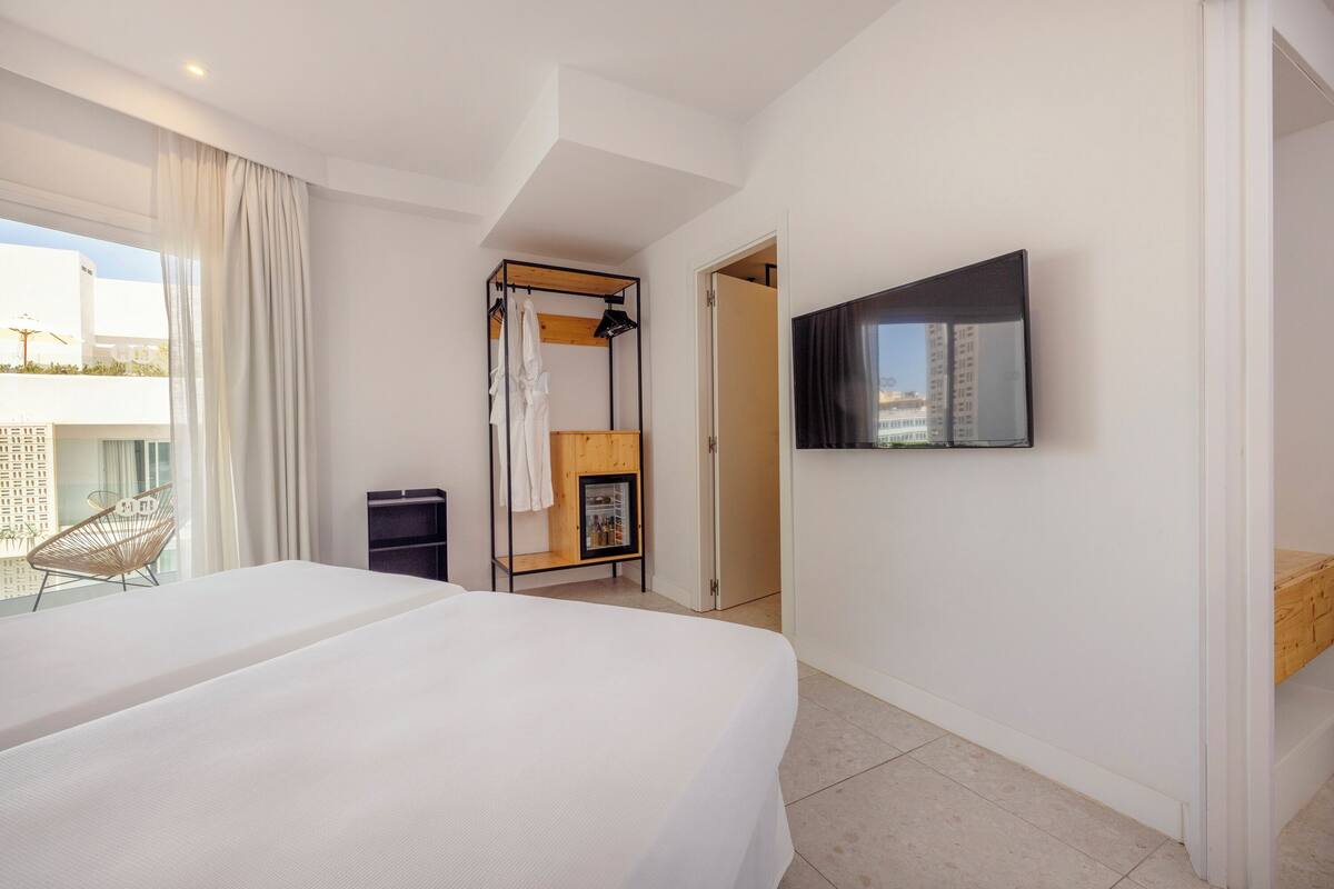 A hotel room with a large bed, a TV mounted on the wall, a wooden shelf with a towel, and a window with curtains.