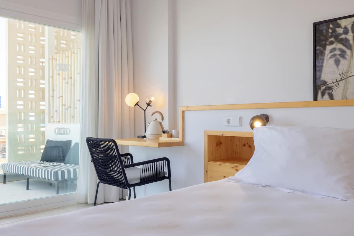 A hotel room with a bed, a desk with a chair, a window with a view of buildings, and a wall-mounted shelf with a lamp.