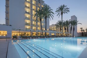 Una piscina con sillas de descanso y sombrillas, rodeada de palmeras y un edificio de hotel con varios pisos y balcones.