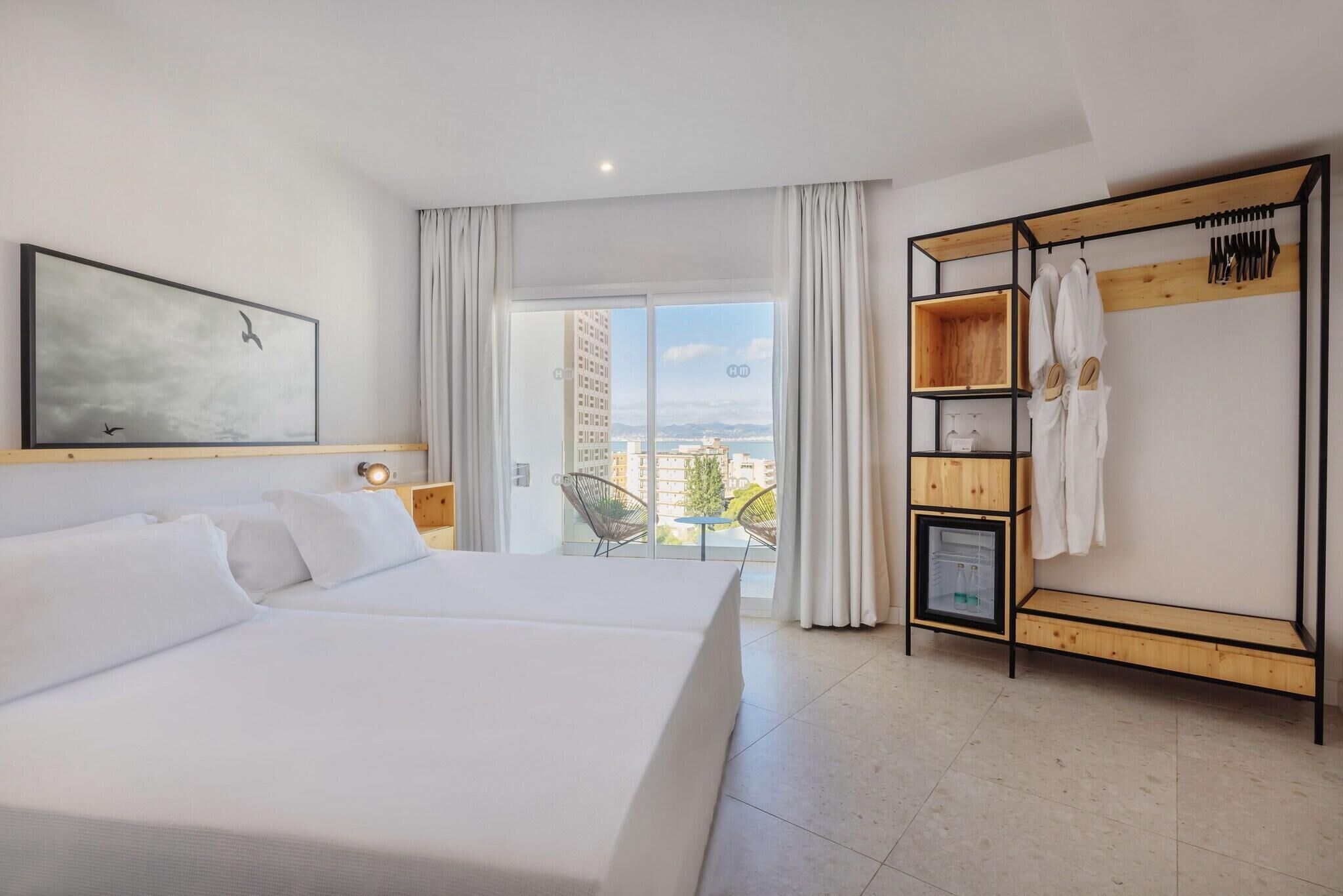 A hotel room with a large bed, a wooden wardrobe, a balcony with a view of buildings, and a painting on the wall.