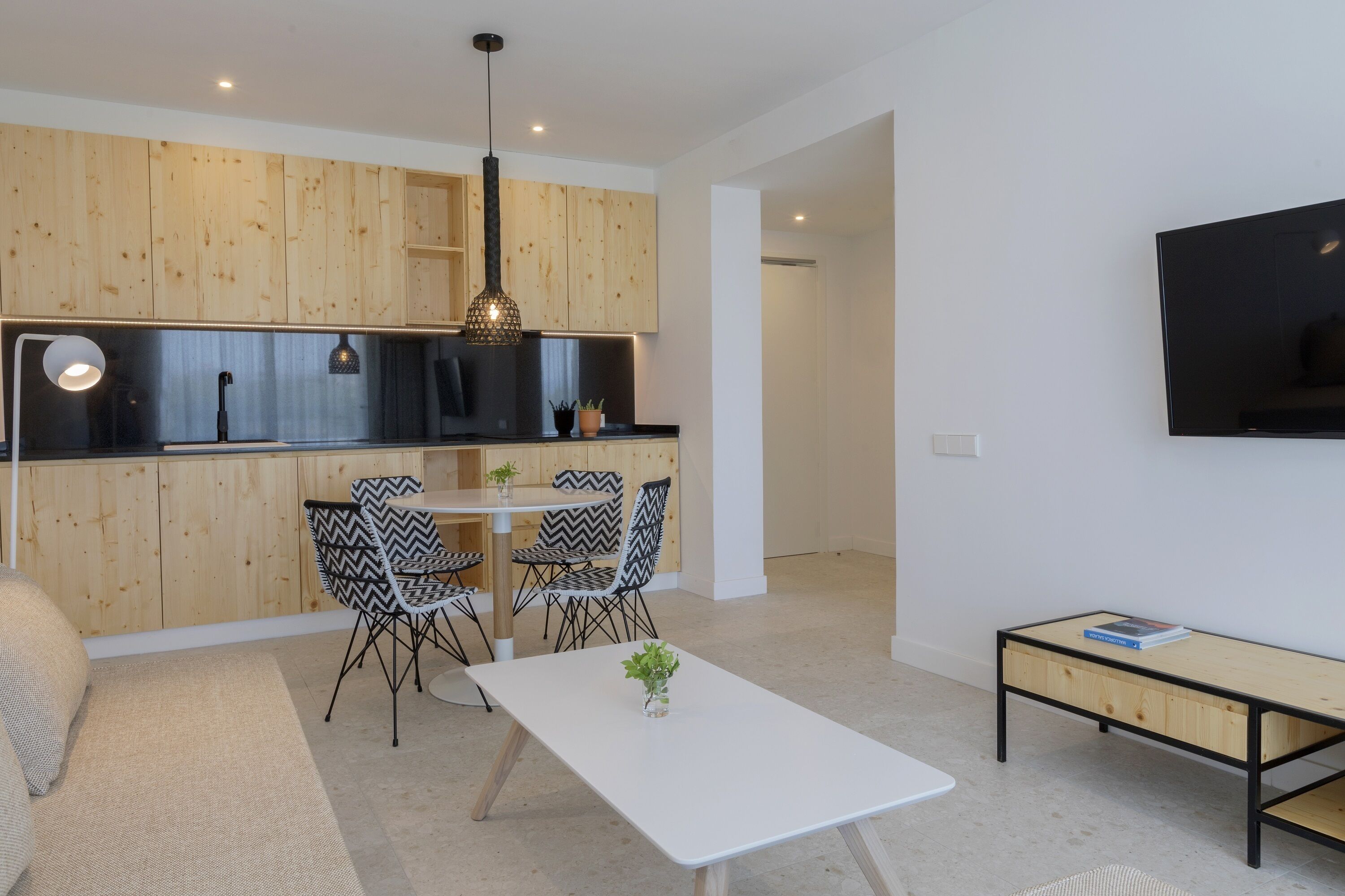 A modern kitchen with wooden cabinets, a dining area with two chairs, a white table, and a TV mounted on the wall.