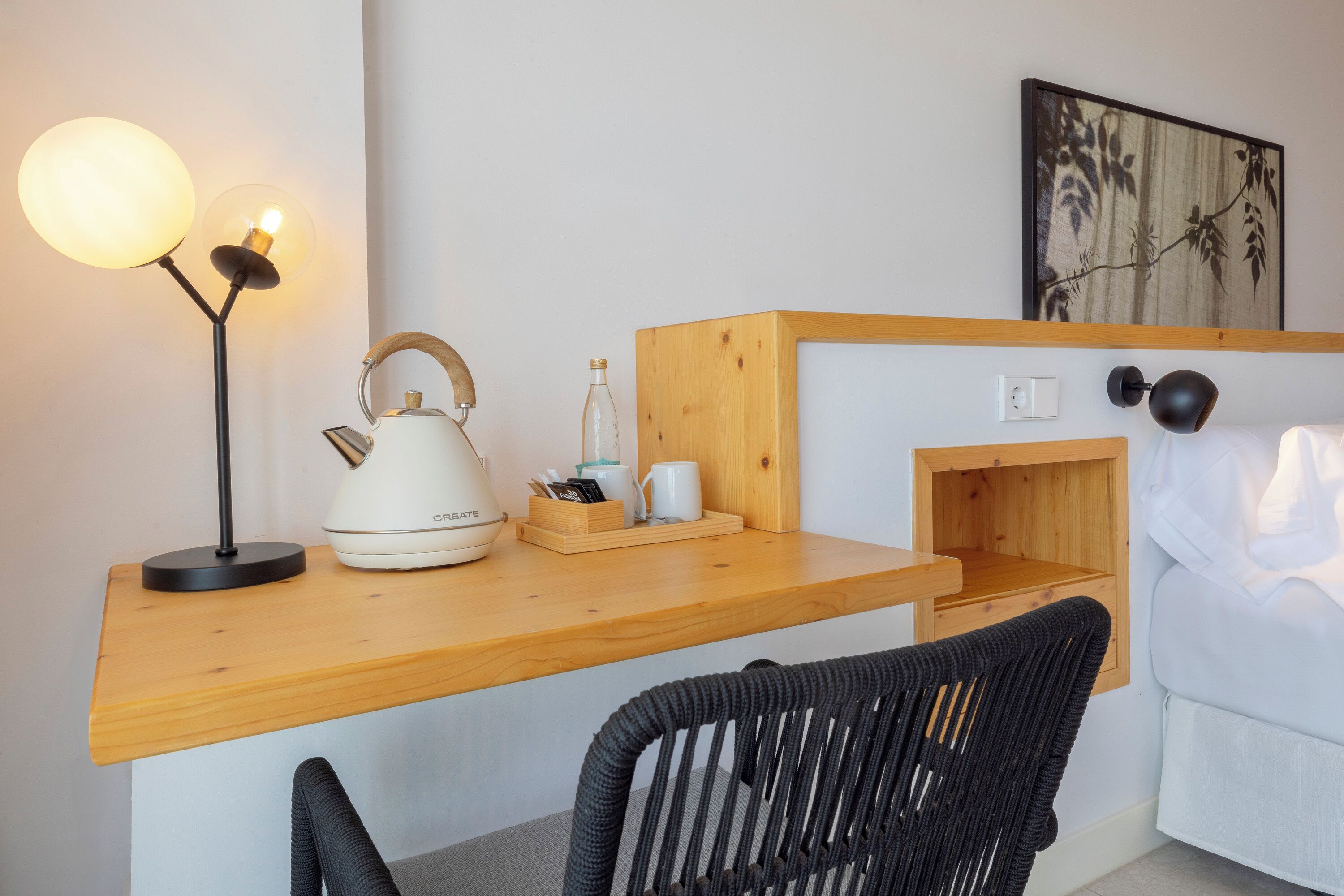 A cozy hotel room with a wooden desk, a kettle, a lamp, and a bed with a white comforter.