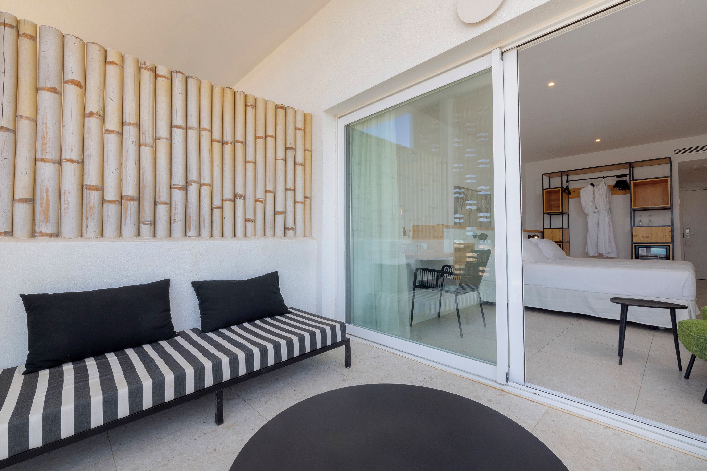 Una habitación de hotel moderna con una puerta corrediza de vidrio que da a un balcón, un sofá a rayas y una cama con una bata blanca colgada en la pared.