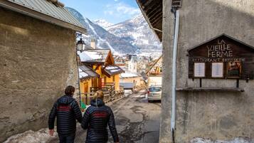 Eine enge Straße mit Häusern, einem Restaurant namens "La Vieille Ferme" und schneebedeckten Bergen im Hintergrund.
