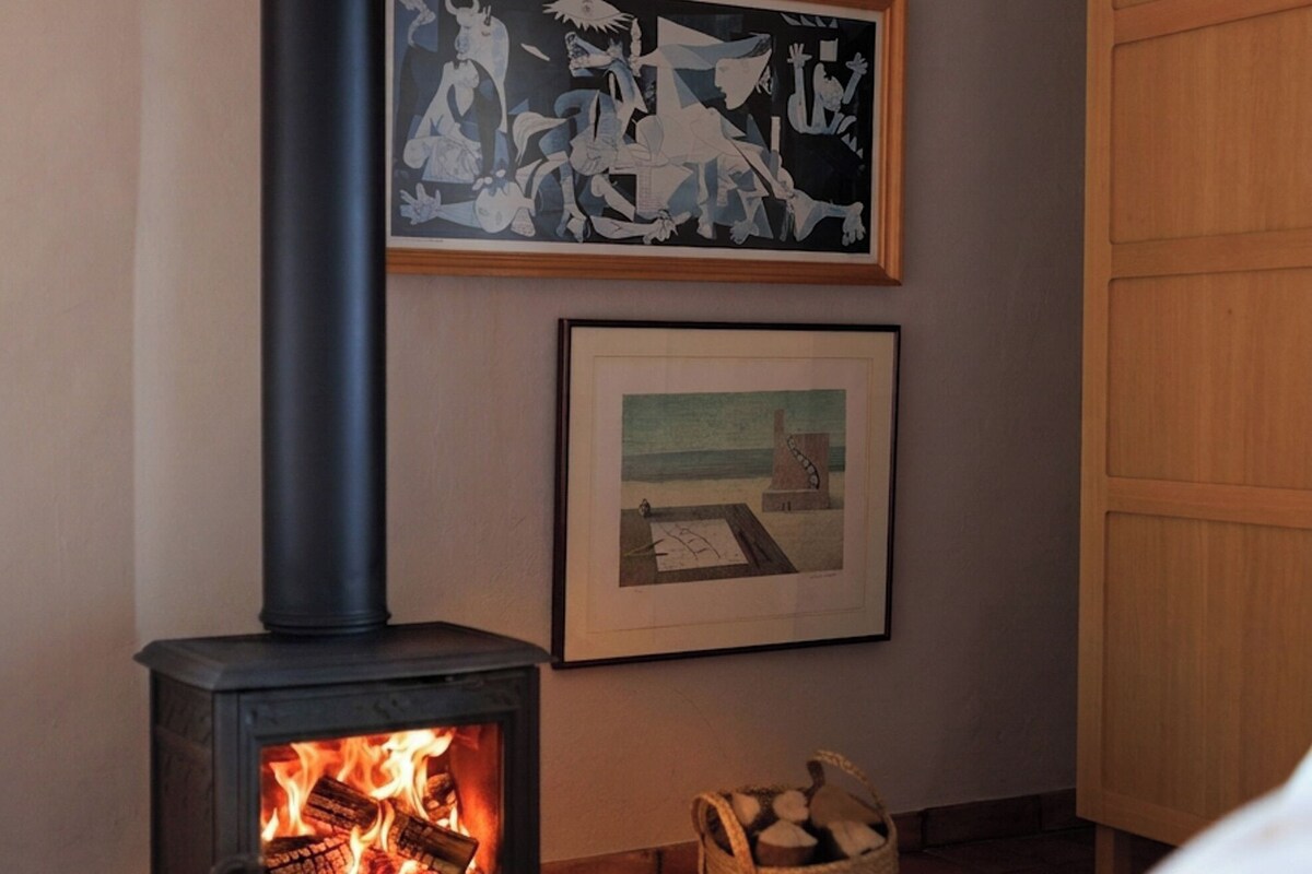 A room with a wood-burning stove, a bed, a basket of firewood, and two framed artworks on the wall.